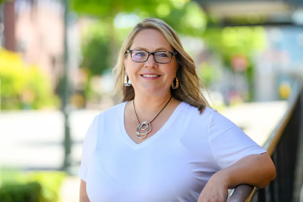 Coach Amy smiling in a white shirt and leaning on a railing outside.