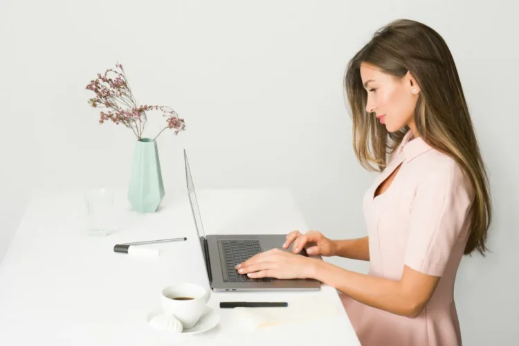 Side profile of woman in pink dress working on a laptop. Grey background, white desk with flowers and cup of coffee