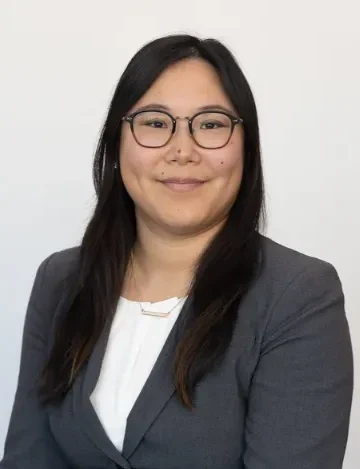 Woman in glasses, long dark hard and grey business jacket smiling for testomonial