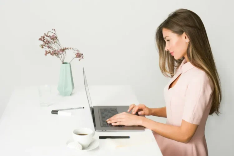 Side profile of woman in pink dress working on a laptop. Grey background, white desk with flowers and cup of coffee