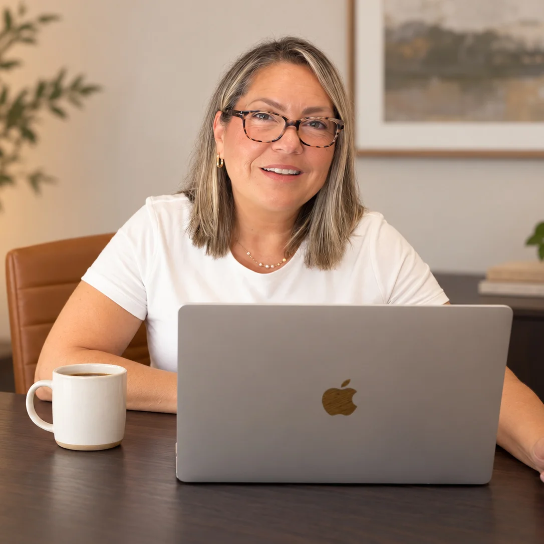 Amy Brainard smiling while seated at a desk with a laptop and coffee mug in a warm, modern office.